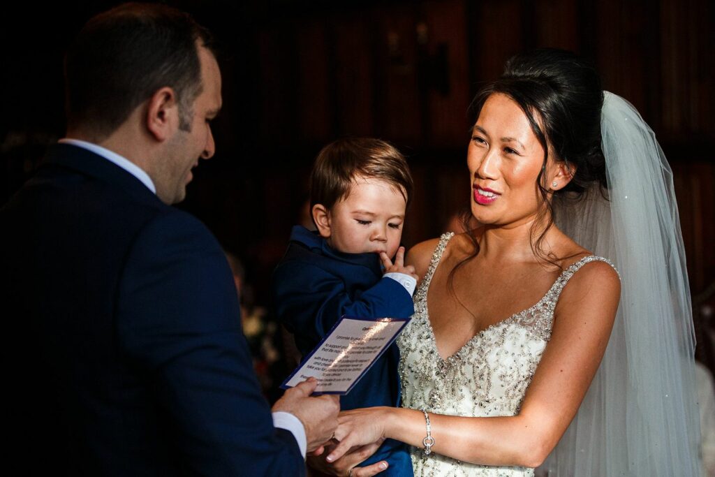 A bride in a white dress, holding a young child, is smiling while facing a groom in a dark suit. The groom is holding a piece of paper, likely reciting vows. The setting appears to be an indoor wedding ceremony. Image by Documentary Wedding Photographer Lewis Bishop Photography.