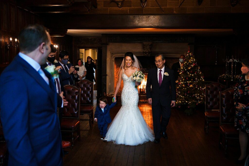 A bride in a white wedding dress walks down the aisle, accompanied by an older man in a suit and tie. They are both smiling. A young boy in a small suit walks beside them. Guests, some of whom are taking photos, watch from the sides. A decorated Christmas tree is visible in the background. Image by Documentary Wedding Photographer Lewis Bishop Photography.