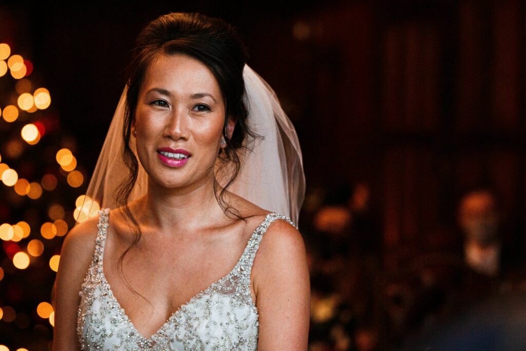 A bride with dark hair in an updo and a sheer white veil stands indoors. She is wearing a white beaded wedding gown and smiling softly, looking slightly to the side. The background is softly lit with blurred, warm-colored lights creating a festive atmosphere. Image by Documentary Wedding Photographer Lewis Bishop Photography.