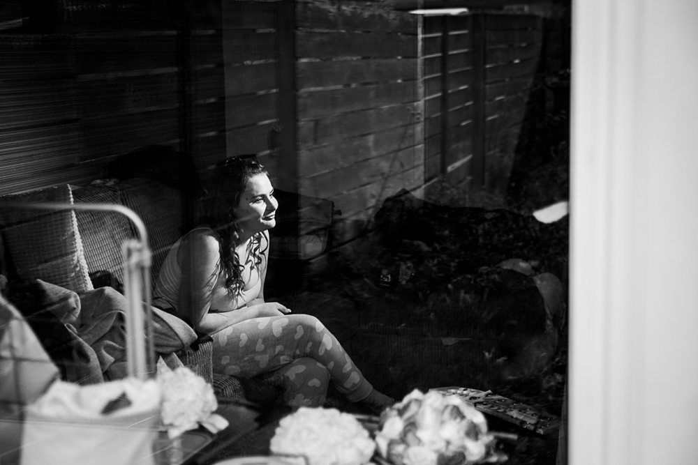 Black and white photo of a woman sitting on a couch visible through a window. She is looking outside and smiling. A blanket is wrapped around her waist, and she is in a relaxed pose. In the foreground, blurred flowers and household items are visible. Image by Documentary Wedding Photographer Lewis Bishop Photography.