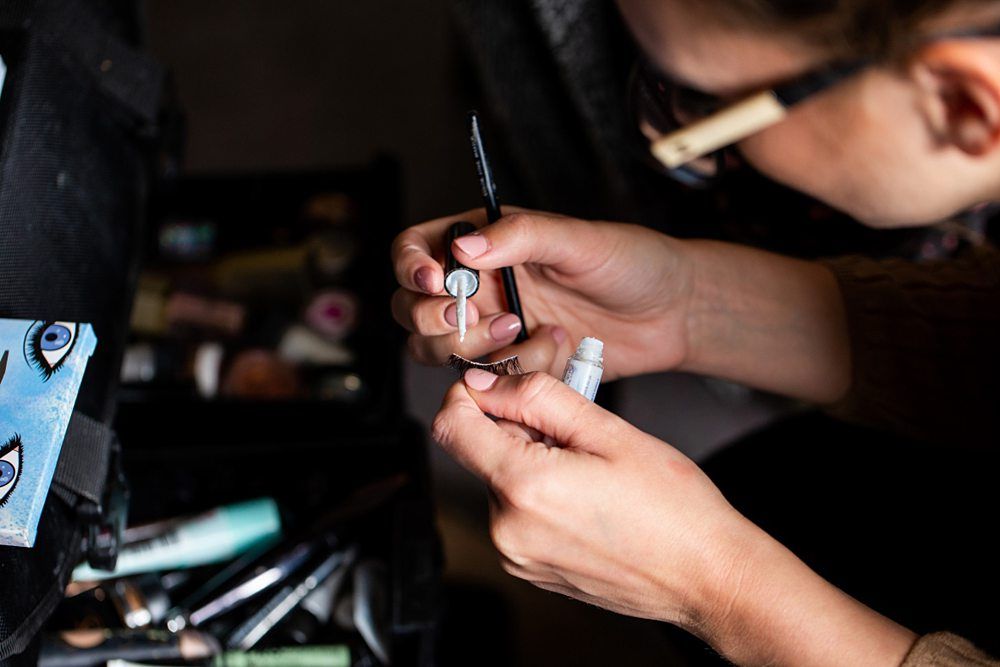 A person applies glue to a false eyelash with tweezers, sitting in front of an open makeup kit containing various beauty products. The person is wearing glasses and the focus is on their hands and the tools they are using. Image by Documentary Wedding Photographer Lewis Bishop Photography.