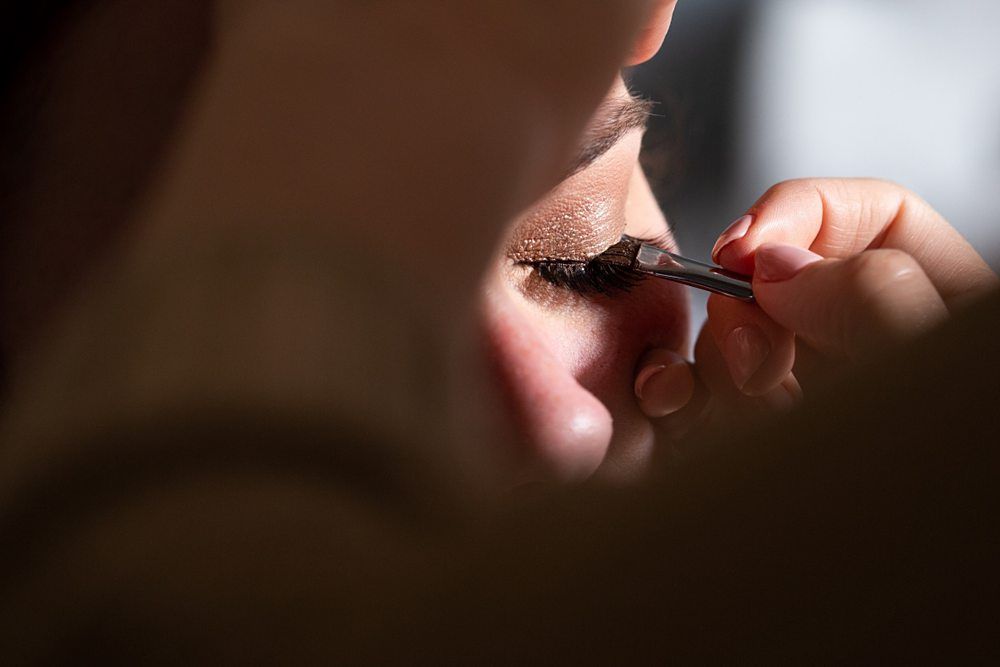 Close-up of a person getting their makeup done, with a focus on the eye. A hand with a brush applies eye shadow or eyeliner to the closed eyelid. The lighting is soft, highlighting the detail of the makeup application. Image by Documentary Wedding Photographer Lewis Bishop Photography.