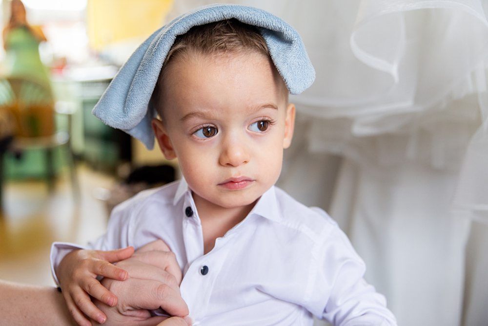 A young child wearing a white button-up shirt has a blue towel draped over their head. The child looks to the side with a pensive expression, while an adult hand gently holds theirs. The background includes blurred elements and another person in the distance. Image by Documentary Wedding Photographer Lewis Bishop Photography.