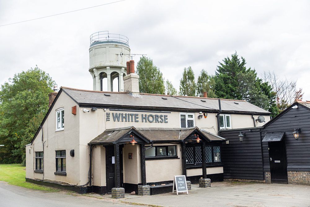 A quaint pub named "The White Horse" with a beige exterior and black roof sits by the roadside. A tall water tower is visible in the background. Trees and greenery surround the area and a small blackboard sign is placed by the entrance. Image by Documentary Wedding Photographer Lewis Bishop Photography.