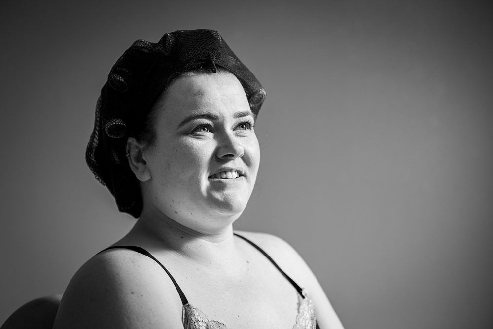 Black and white photo of a person smiling and looking slightly away from the camera. They are wearing a hair net and seated against a plain background, dressed in a strappy top. The lighting is soft, giving the image a calm and serene atmosphere. Image by Documentary Wedding Photographer Lewis Bishop Photography.