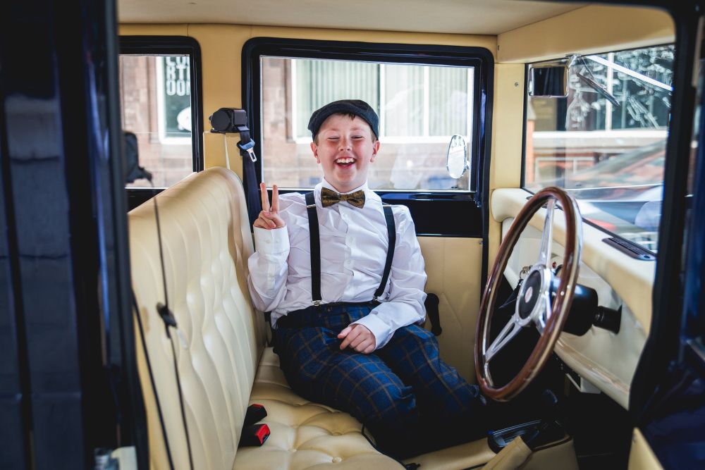 A young boy in vintage attire, including a bow tie, suspenders, and a cap, is sitting in the back seat of a classic car. He is smiling widely and flashing a peace sign with his right hand. The car's interior features cream-colored upholstery. Image by Documentary Wedding Photographer Lewis Bishop Photography.