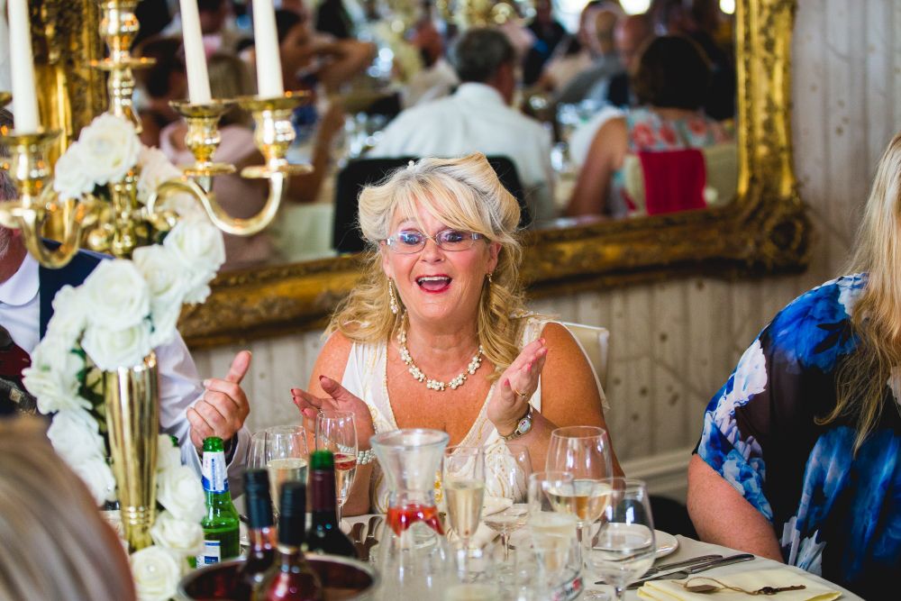 A woman with blonde hair and glasses, dressed in a light-colored outfit, sits at an elegantly set table, clapping her hands. She is surrounded by other people who are engaged in conversation. The table has white flowers, candles, and various drinks. Image by Documentary Wedding Photographer Lewis Bishop Photography.