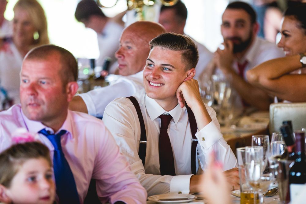 A young man in a white shirt and maroon tie sits at a table, smiling and resting his chin on his hand during a lively gathering. People around him are engaged in conversation, featuring glasses of drinks and table decorations. Image by Documentary Wedding Photographer Lewis Bishop Photography.