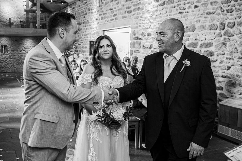 A man in a suit shakes hands with another man in a suit at a wedding ceremony, with a woman in a white dress standing between them. Stone wall and seated guests are in the background. Image by Documentary Wedding Photographer Lewis Bishop Photography.