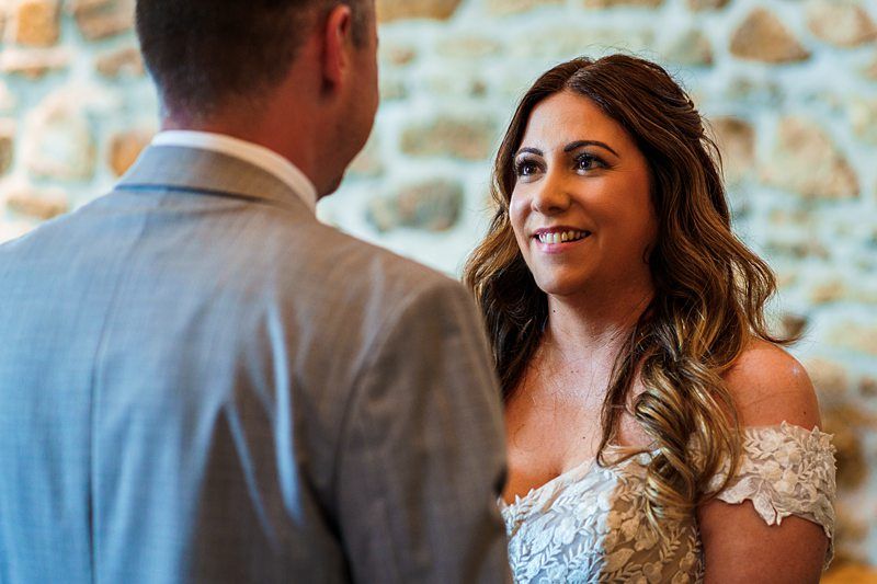 A bride wearing a white dress smiles at a groom in a gray suit; they are standing against a stone wall background. Image by Documentary Wedding Photographer Lewis Bishop Photography.