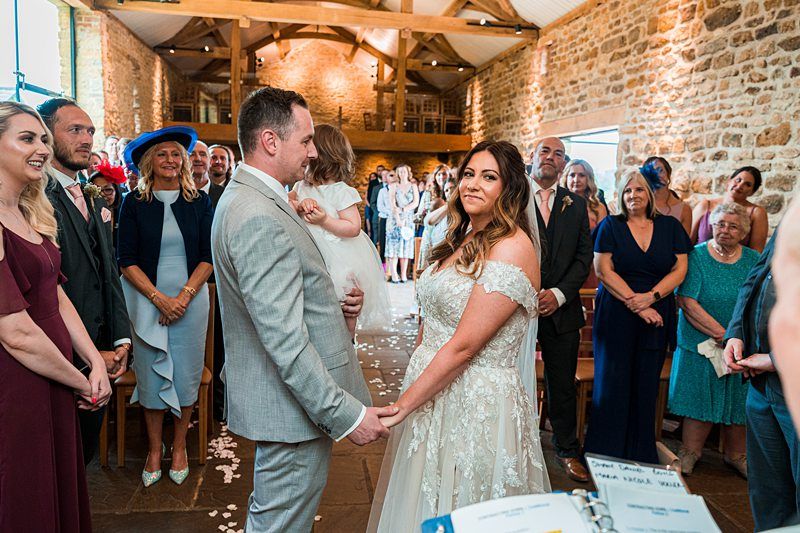 A bride and groom stand facing each other, holding hands, during their wedding ceremony in a rustic indoor setting. Guests are seated and standing around them, watching the couple. Image by Documentary Wedding Photographer Lewis Bishop Photography.