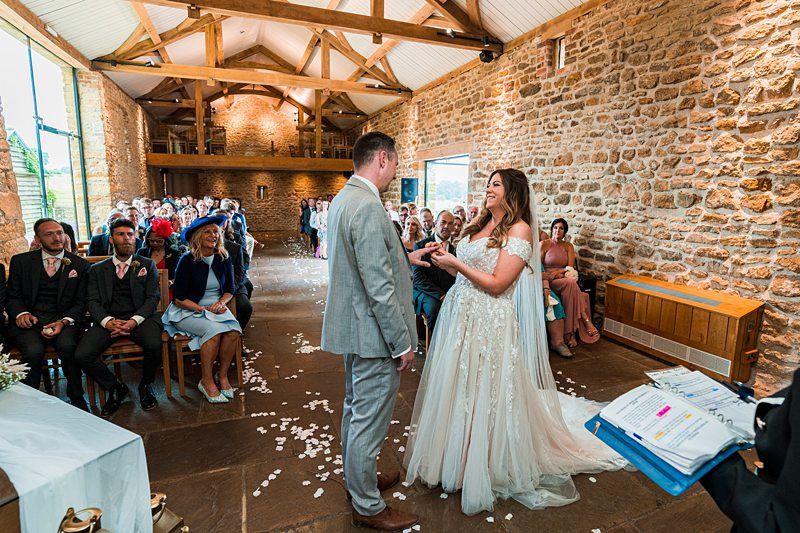 A bride and groom stand facing each other in a rustic, stone-walled venue with seated guests watching their wedding ceremony. The bride is in a white gown, and the groom is in a light gray suit. Image by Documentary Wedding Photographer Lewis Bishop Photography.