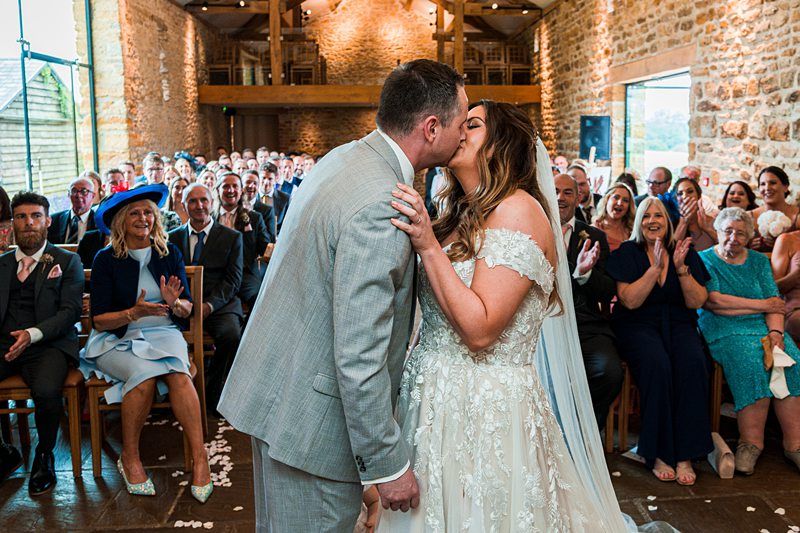 A couple in wedding attire kisses at their indoor ceremony while guests sit and watch, many smiling and clapping, in a rustic venue with stone walls and wooden beams. Image by Documentary Wedding Photographer Lewis Bishop Photography.