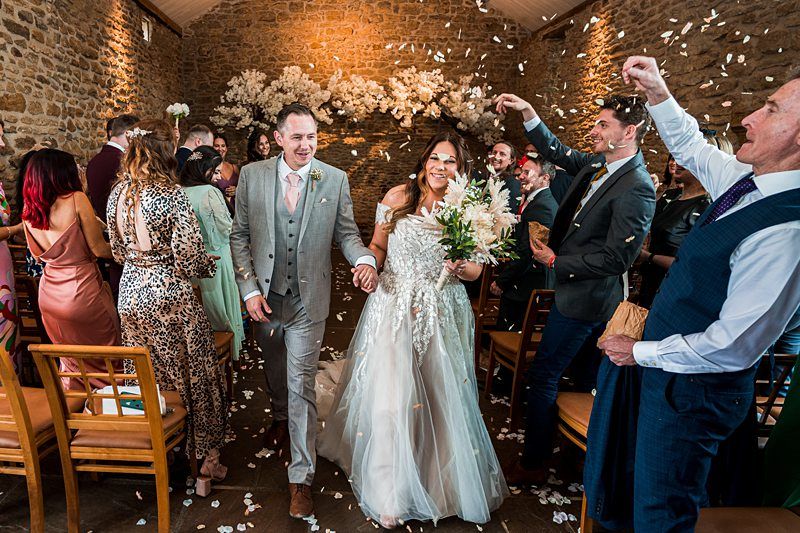 A bride and groom walk down the aisle as guests throw flower petals in celebration. The scene is indoors with a stone wall backdrop and floral decorations. Image by Documentary Wedding Photographer Lewis Bishop Photography.