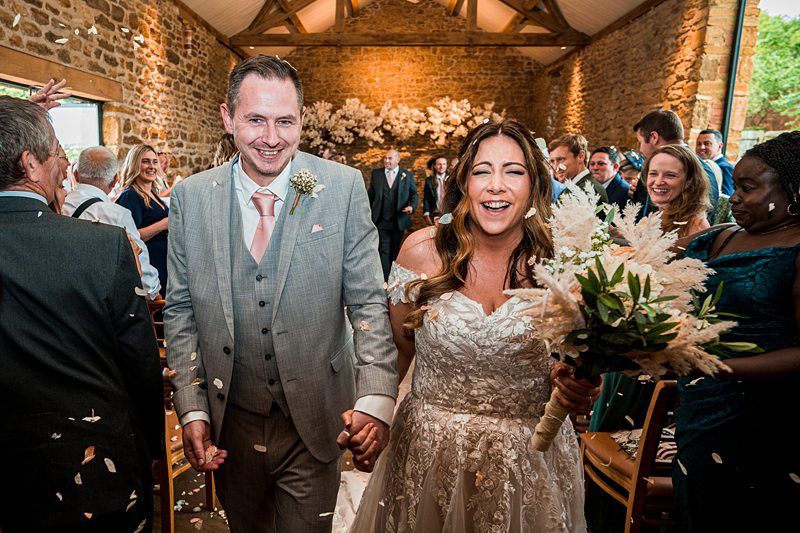 A bride and groom smile and hold hands while walking down the aisle as guests throw flower petals. The setting is a rustic indoor venue with brick walls and a floral arrangement backdrop. Image by Documentary Wedding Photographer Lewis Bishop Photography.