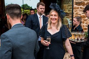 A woman in a black dress and large black fascinator holds a glass of champagne at an outdoor event. She is among other formally dressed people, with a waiter holding a tray of drinks in the background. Image by Documentary Wedding Photographer Lewis Bishop Photography.