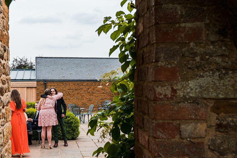 Three people are seen on a patio area outside a stone building. One person in a pink dress hugs a person in dark clothing, while another person in an orange dress stands nearby. Image by Documentary Wedding Photographer Lewis Bishop Photography.