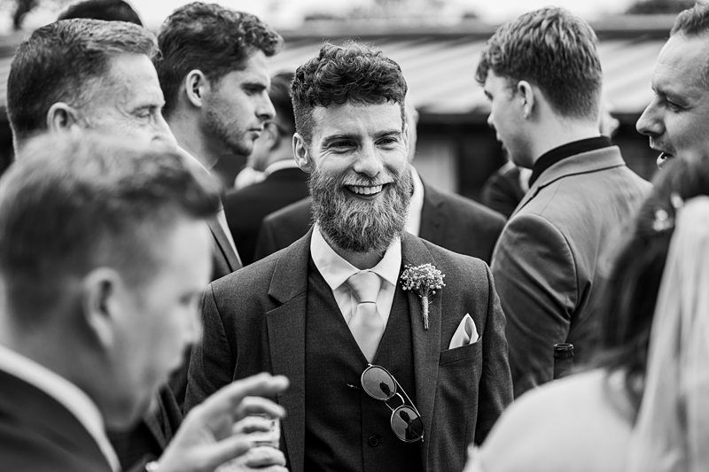 A man with a beard and sunglasses hanging from his suit smiles while standing among a group of people dressed in formal attire. Image by Documentary Wedding Photographer Lewis Bishop Photography.