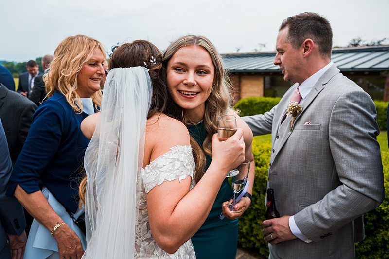 A bride in a white dress and veil hugs a woman holding a glass of wine, while people in formal attire converse in the background at an outdoor gathering. Image by Documentary Wedding Photographer Lewis Bishop Photography.