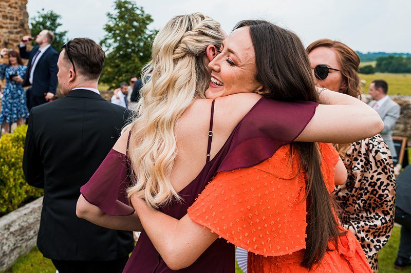 Two women embracing warmly at an outdoor gathering, with other people and greenery visible in the background. Image by Documentary Wedding Photographer Lewis Bishop Photography.