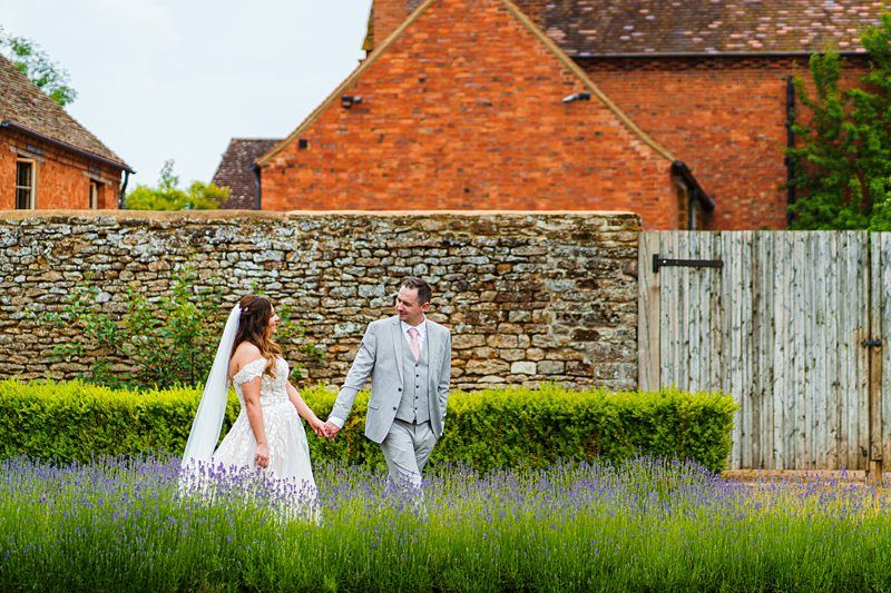 A couple in wedding attire holds hands while walking through a lavender garden in front of a brick wall and rustic buildings. Image by Documentary Wedding Photographer Lewis Bishop Photography.