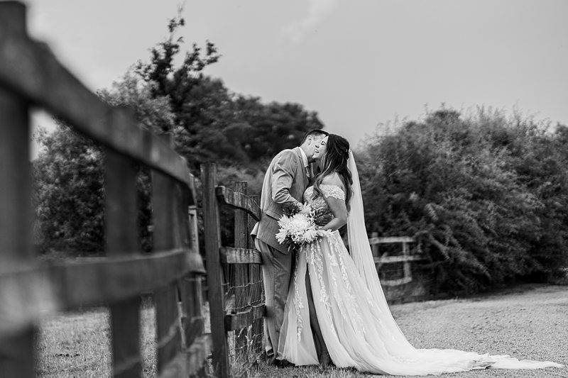 A newlywed couple shares a kiss by a wooden fence in a countryside setting. The bride holds a bouquet and wears a long, flowing dress with a veil. The groom wears a suit. Black and white photo. Image by Documentary Wedding Photographer Lewis Bishop Photography.
