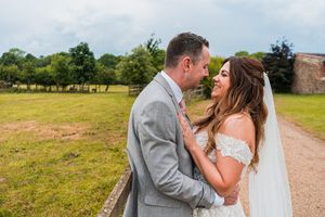 A bride in a white wedding dress and a groom in a gray suit stand outdoors, smiling and looking at each other, with greenery and trees in the background. Image by Documentary Wedding Photographer Lewis Bishop Photography.