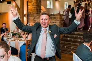 A man in a suit and tie raises both arms in the air, smiling joyfully, at a formal indoor gathering. People are seated at tables in the background. Image by Documentary Wedding Photographer Lewis Bishop Photography.