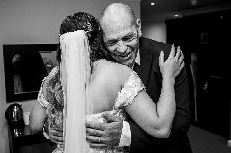 A bride and an older man share an embrace in a room. The woman is wearing a wedding dress with a veil, and the man is smiling with his eyes closed. The setting appears to be indoors. Image by Documentary Wedding Photographer Lewis Bishop Photography.