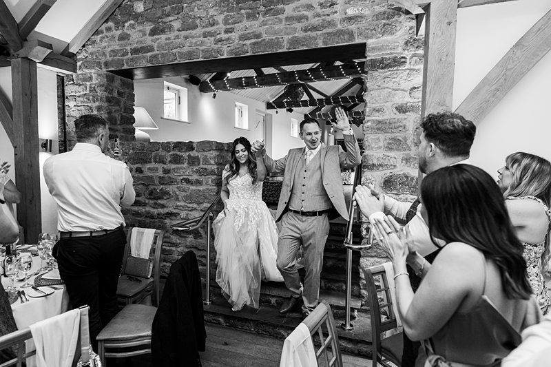 A bride and groom enter a reception area while guests clap and stand in applause. The setting features rustic stone walls and wooden beams. Image by Documentary Wedding Photographer Lewis Bishop Photography.