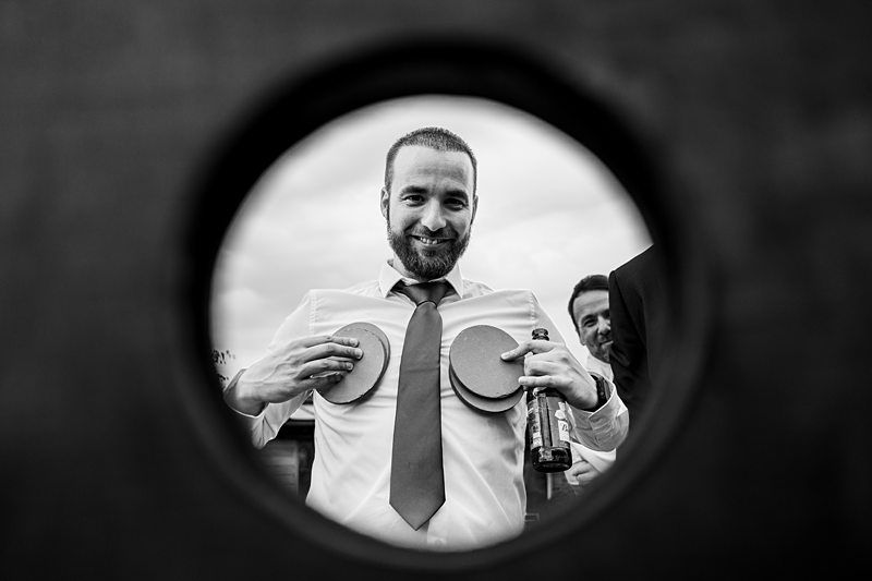 A man wearing a tie poses playfully with two round objects held to his chest, viewed through a circular frame. He smiles at the camera, while another person is partially visible in the background. Image by Documentary Wedding Photographer Lewis Bishop Photography.