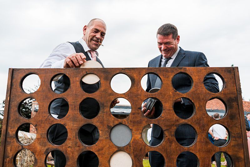Two men in suits are playing a large outdoor wooden Connect Four game. One man holds a game piece while the other places one in a slot. Image by Documentary Wedding Photographer Lewis Bishop Photography.