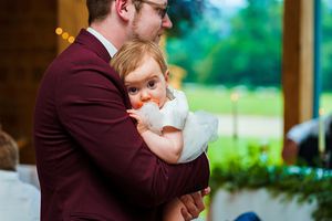 A person in a burgundy suit holds a baby dressed in white, who appears to be biting an orange pacifier. Image by Documentary Wedding Photographer Lewis Bishop Photography.