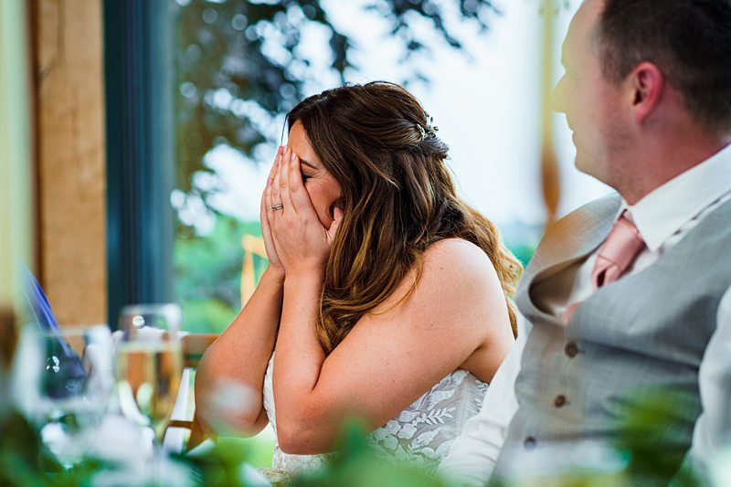 A woman sits at a table, covering her face with her hands. A man in a vest and tie sits beside her, looking in her direction. The background shows some greenery and a window. Image by Documentary Wedding Photographer Lewis Bishop Photography.