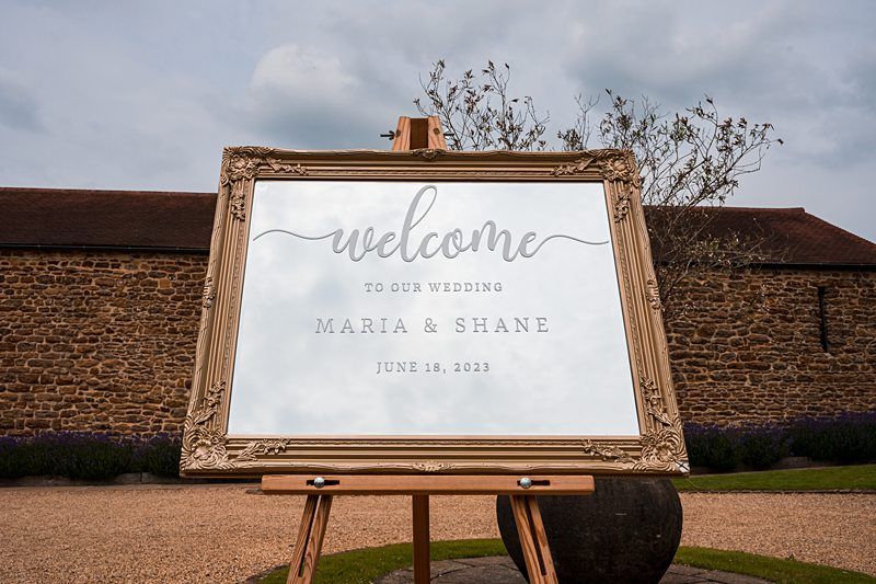 A decorative sign on an easel reads, "Welcome to our wedding, Maria & Shane, June 18, 2023" with a rustic building in the background. Image by Documentary Wedding Photographer Lewis Bishop Photography.