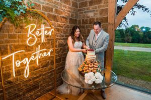 A bride and groom cut a wedding cake on a glass table with donuts and flowers, next to a neon sign reading "Better Together." They are in a rustic venue with a view of greenery through a large window. Image by Documentary Wedding Photographer Lewis Bishop Photography.