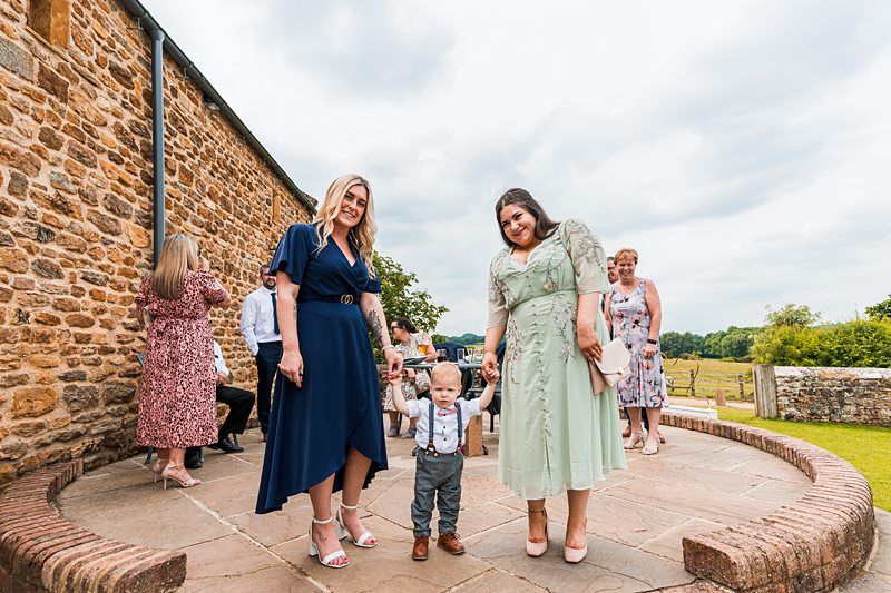 Two women in dresses and a small child stand outdoors near a stone building, with other people visible in the background on a cloudy day. Image by Documentary Wedding Photographer Lewis Bishop Photography.