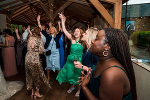 A group of people are dancing energetically at an indoor event, with arms raised and expressions of joy on their faces. The venue features wooden beams and a large window. Image by Documentary Wedding Photographer Lewis Bishop Photography.