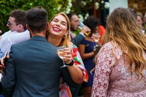 A woman holding a glass of wine smiles while embracing a man at an outdoor gathering, with other people socializing in the background. Image by Documentary Wedding Photographer Lewis Bishop Photography.
