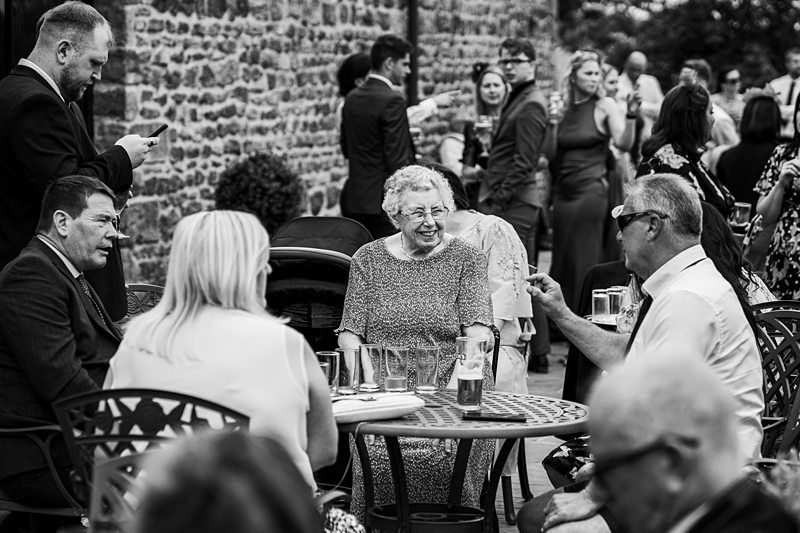 A group of people, including an elderly woman, sit and stand outside a stone building, engaging in conversation and using mobile phones. Image by Documentary Wedding Photographer Lewis Bishop Photography.