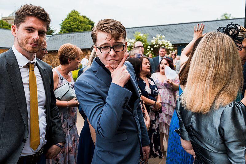 Group of people at an outdoor event. A young man in a blue suit and glasses poses with his hand on his chin, looking at the camera. Others around him are engaged in conversation. Image by Documentary Wedding Photographer Lewis Bishop Photography.