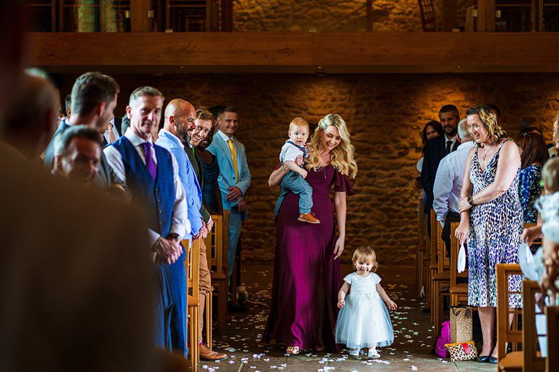 A woman in a purple dress walks down the aisle holding a baby while leading a young girl in a white dress at a wedding ceremony. Guests seated along the aisle smile and watch. Image by Documentary Wedding Photographer Lewis Bishop Photography.