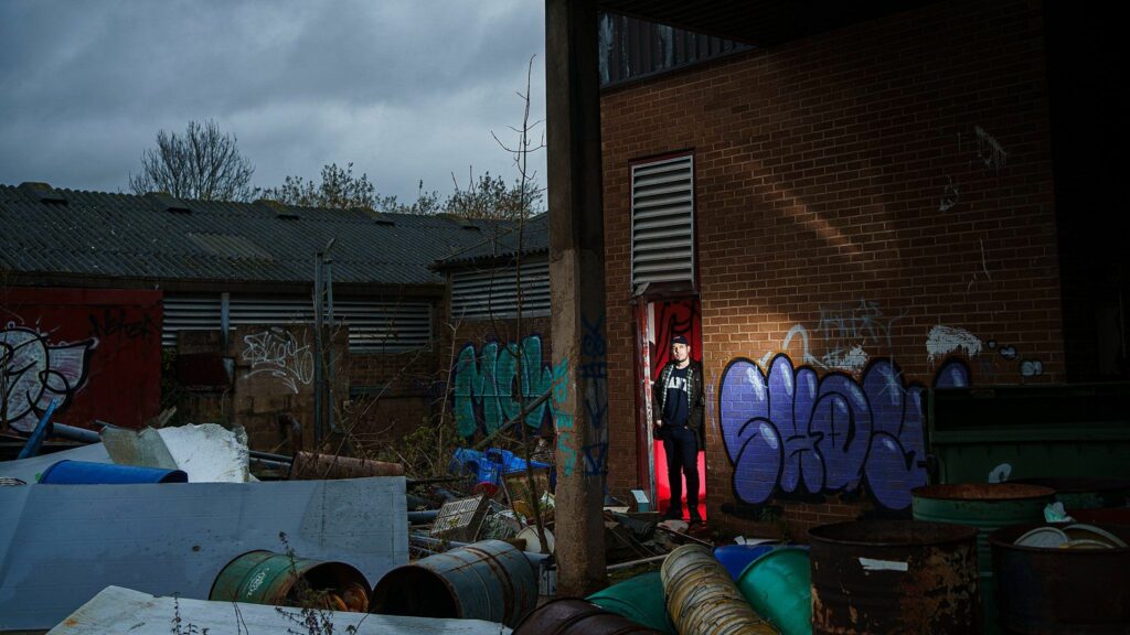 A lone person wearing a dark hoodie and red pants stands in the doorway of a dilapidated brick building covered in graffiti. Surrounding the building are scattered debris and rusted metal drums, set against a gloomy, overcast sky. Image by Documentary Wedding Photographer Lewis Bishop Photography.