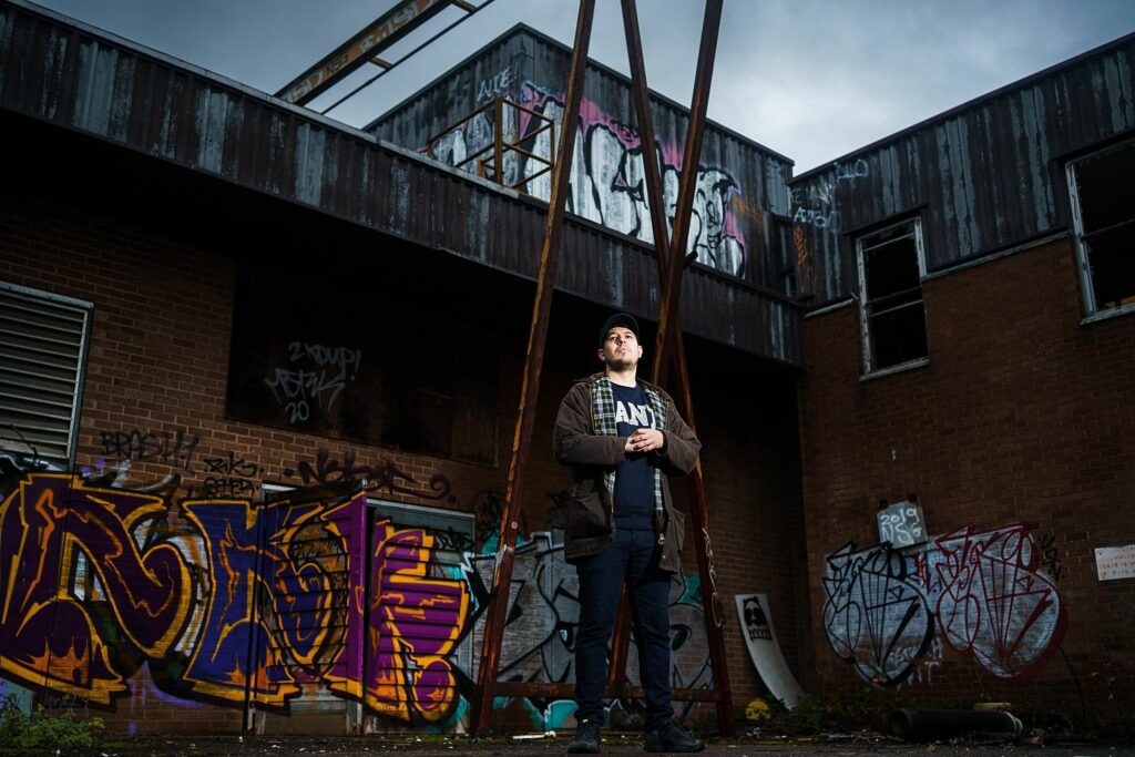 A person stands in a dark outdoor space with brick buildings covered in colorful graffiti in the background. They wear a cap, dark jacket, and jeans, looking upwards with hands in pockets. The sky is cloudy, and the scene feels moody and urban. Image by Documentary Wedding Photographer Lewis Bishop Photography.