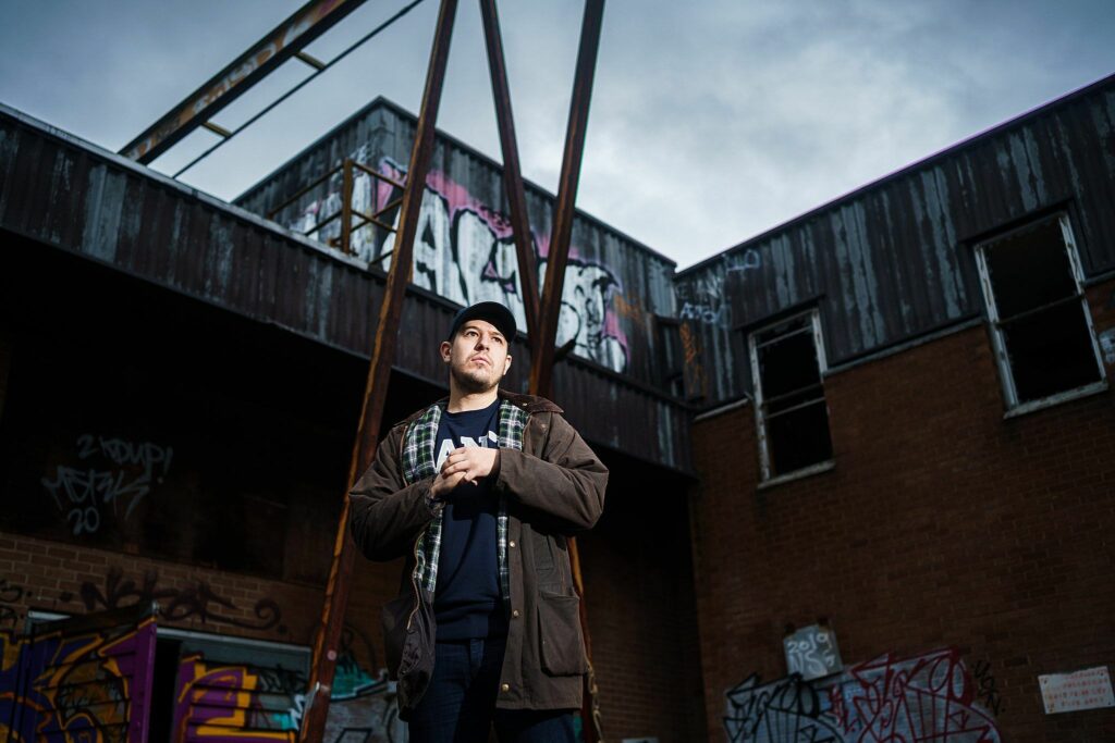 A man stands outdoors wearing a dark jacket and cap, with his hands clasped in front of him. The backdrop features an aged, graffiti-covered brick building and a cloudy sky overhead, creating an urban, moody atmosphere. Image by Documentary Wedding Photographer Lewis Bishop Photography.