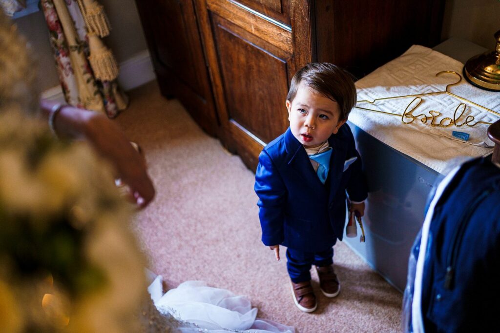 A young boy in a blue suit, holding an object in his left hand, looks up with a curious expression. He stands indoors near a wooden cabinet and a table with a hanger labeled "bride." A wedding dress and decorations are visible around him. Image by Documentary Wedding Photographer Lewis Bishop Photography.