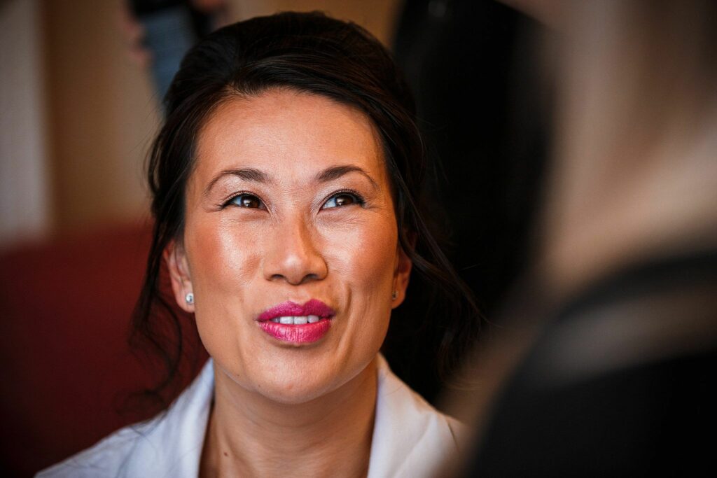 A person with dark hair, styled up, is smiling and looking slightly upwards. They have bright pink lipstick, subtle eye makeup, and are wearing a white shirt. The background is blurred with a hint of another person in the foreground. Image by Documentary Wedding Photographer Lewis Bishop Photography.