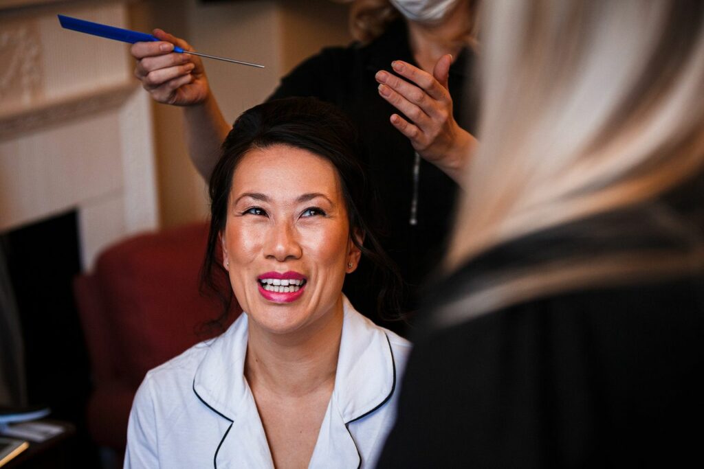 A smiling woman with dark hair, wearing a white top, is getting her hair styled by a person blurred in the foreground and holding a blue comb. The setting appears to be indoors, perhaps in a living room, with a red chair and a fireplace in the background. Image by Documentary Wedding Photographer Lewis Bishop Photography.