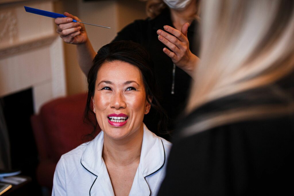 A woman, dressed in a white top, smiles joyfully as a hairstylist works on her hair. Another person wearing a mask and black attire assists in the background. She is seated in a well-lit room with red and white furnishings. Image by Documentary Wedding Photographer Lewis Bishop Photography.