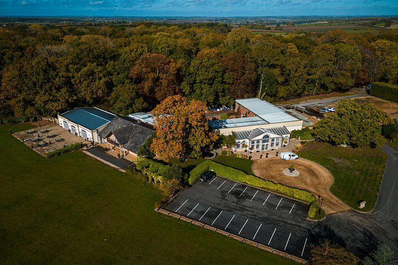 Aerial view of a countryside facility surrounded by dense trees. The property includes several connected buildings, open areas, and a small parking lot with a few cars. A circular drive with a central feature sits in front of the building entrance. Image by Documentary Wedding Photographer Lewis Bishop Photography.